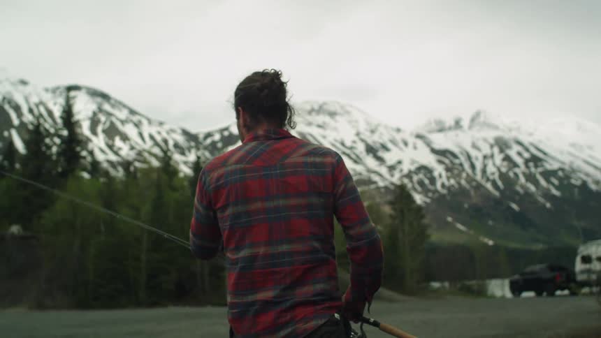 A man in a flannel shirt grabs the fishing line of his fishing rod as he walks through a forest clearing