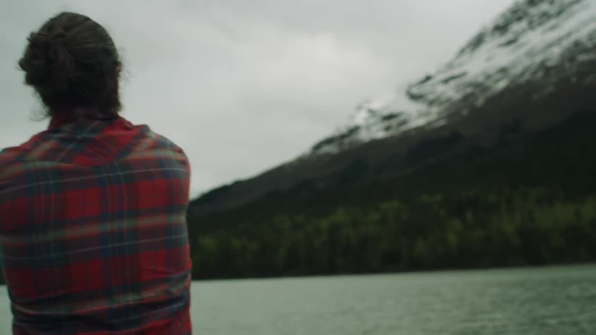 Man standing on shore casts fishing line with snow covered mountains across water under overcast sky