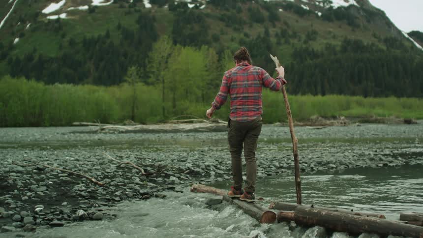 A man uses a long stick to help him balance as he crosses a stream using rocks and logs