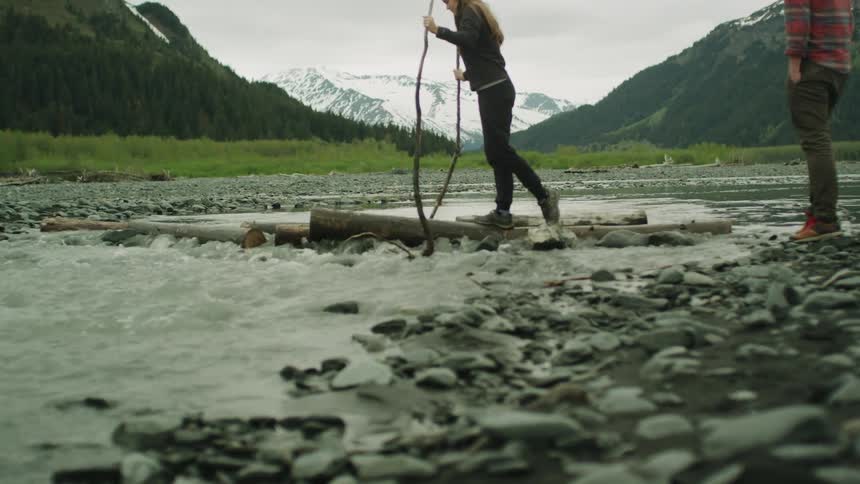 Man watches woman trying to cross river using fallen logs and sticks with mountains and forest in background