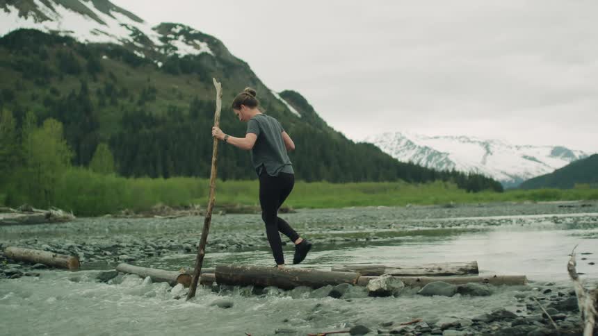 A woman uses a long branch to keep her balance while walking across a row of logs spanning a river in the wilderness on a cloudy day