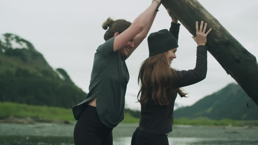 Two women pushing a log with a mountain range the sky and clouds in the background