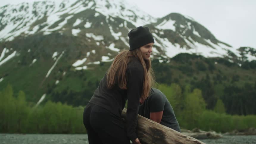 A woman runs to another womans side to help lift a large log in the wilderness on a cloudy day