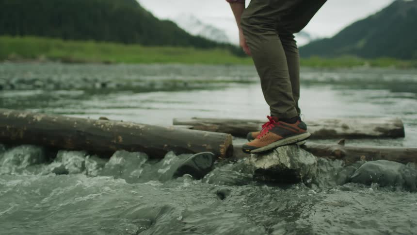 A man balances on a rock and then reaches down to touch a log and get his balance as he crosses a river