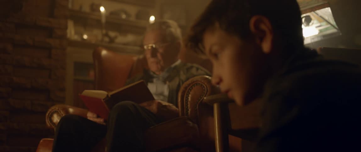 A boy plays at the feet of his grandfather that is reading a book out loud