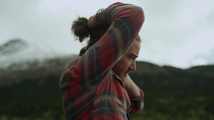 A man stands at the base of a mountain and pulls his hair into a pony tail