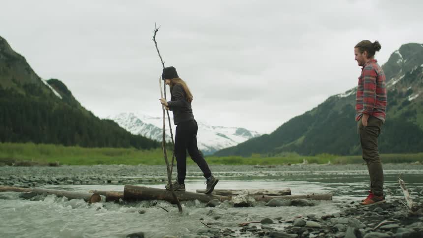 Couple crossing a shallow river over logs and rocks near the mountain
