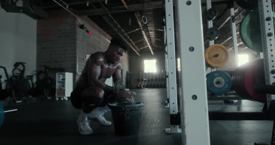 A shirtless man kneels before a bucket and uses the chalk within to cover his palms