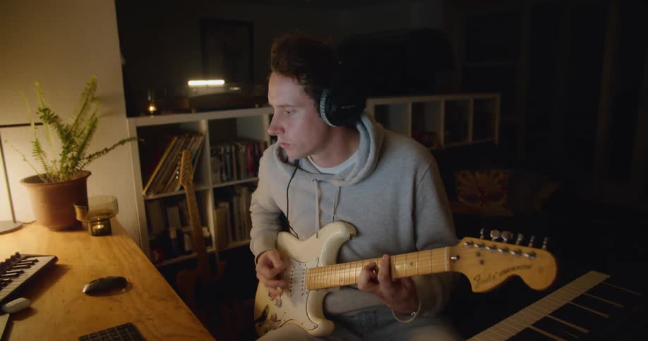 A man strums his guitar while looking at a laptop