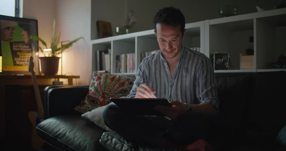 Man sits cross legged on sofa sketching on tablet and smiling