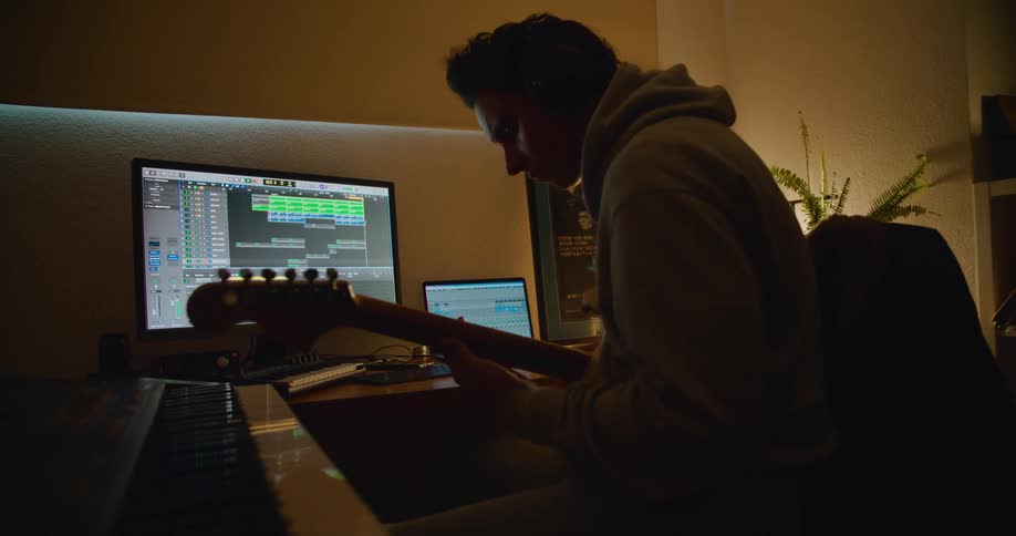 Man with gray hoodie plays guitar looking at desktop and laptop