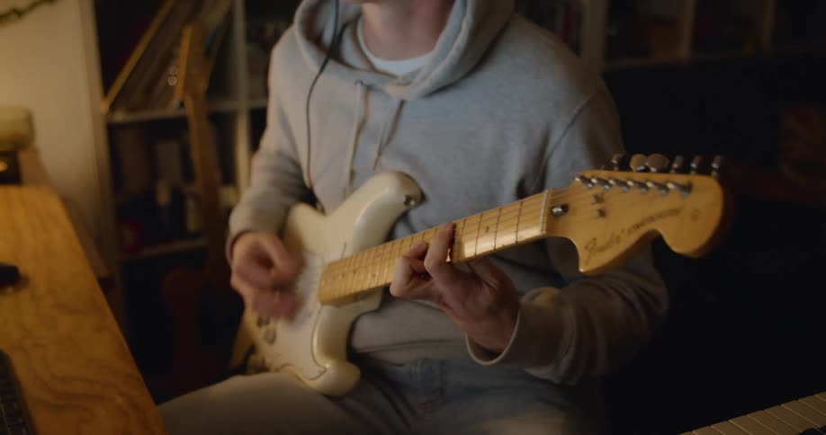 A man wearing headphones plays his guitar while sitting at a desk in a dimly lit room