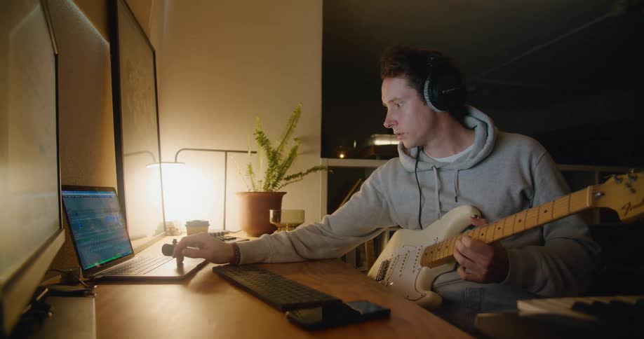 A man holds a guitar and makes adjustments on a laptop before starting to play the guitar