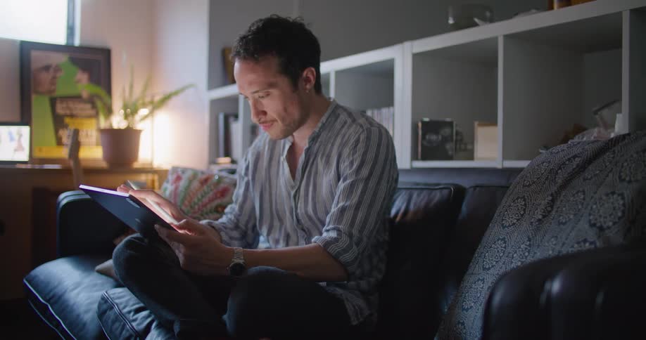 Man with shirts and jeans sitting on black couch and using tablet smiling