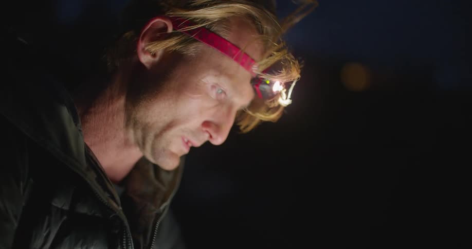 A man wearing a headlamp is busy working on a smartphone while outdoors on a cool and windy night