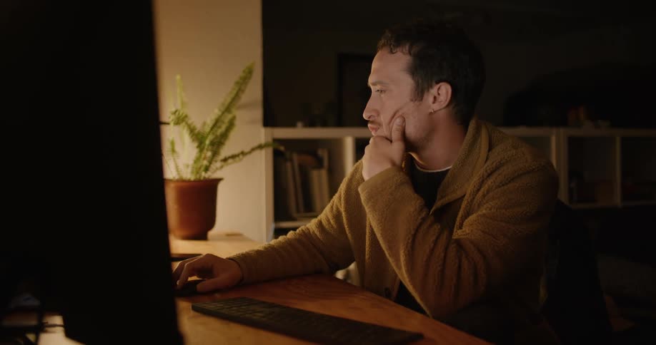Man sits at table working on computer in dark room lit by lamp