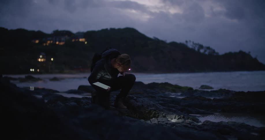 A man types something on his tablet as he crouches on a beach