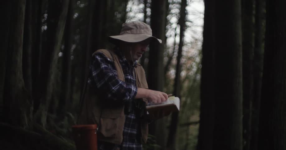 A man standing in the forest takes out a notebook and pencil while bird watching