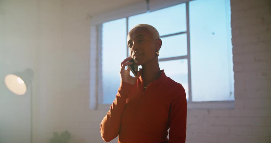 A woman smiles and laughs as she stands talking on the phone near a window in a bright sunny room