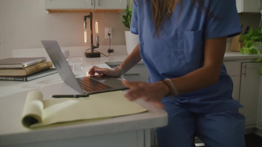 A woman wearing scrubs walks into a kitchen with a laptop and sits down