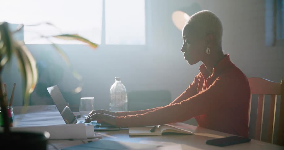 A woman in a room writing in a notebook and using a laptop with a wall and window in the background