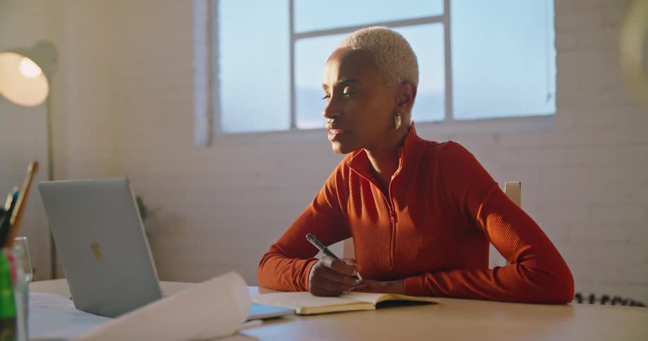 A young woman writes in a notebook and waves at her laptop camera while participating in a video conference