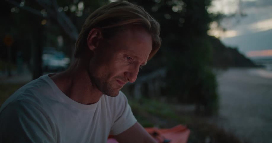A man rubs his face and cries as he looks at his phone sitting next to a kayak overlooking the beach