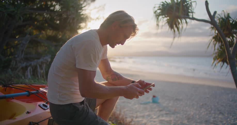 A man kneeling on a sandy beach next to a kayak while holding a cell phone