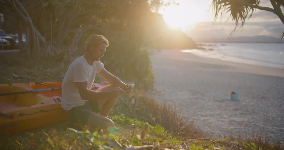 Blonde man with white t shirt and black shorts squatting down next to yellow and red boat takes photos of beach and sea on smartphone at the beach
