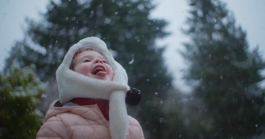 Blonde girl with white hat pink coat opens her mouth while snowing in the woods