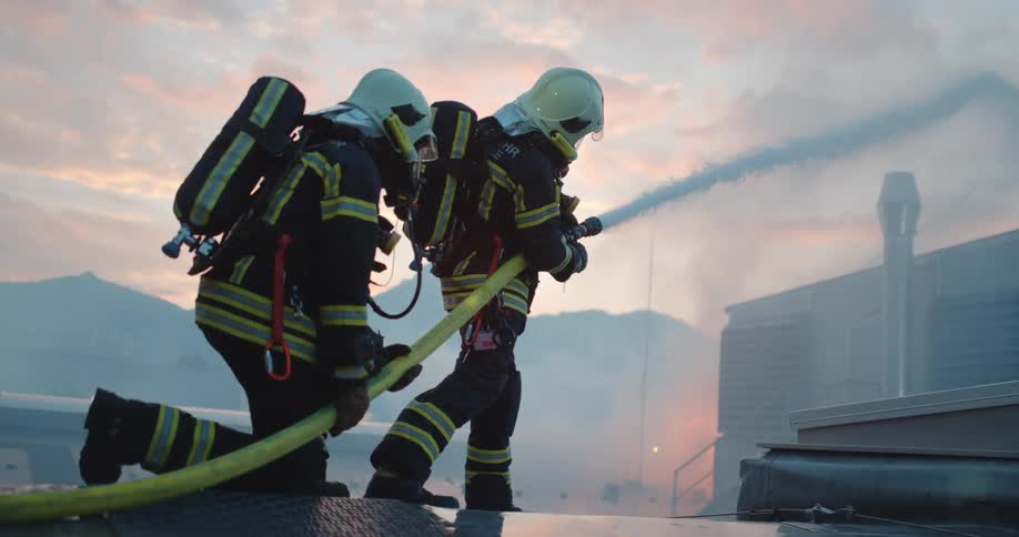 Two firefighters with helmets spraying a water hose with the sky and clouds in the background