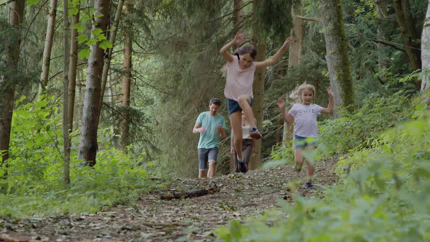 Parents walk along trail through wood in unison as daughters run and jump ahead