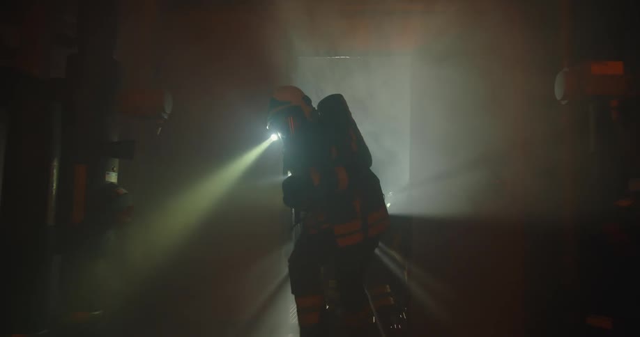 Two firefighters with flashlights open a door to enter a smoke filled room