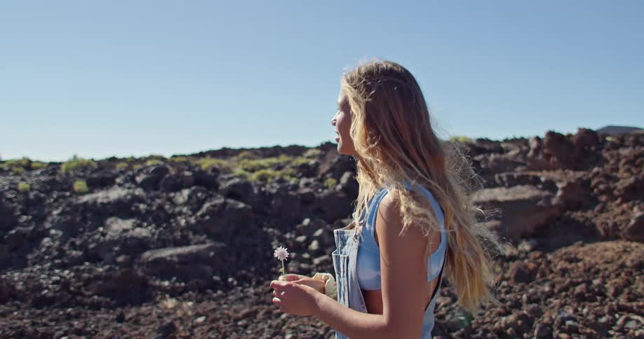 A woman with a flower in her hand walking with a rocky landscape the sky and clouds in the background