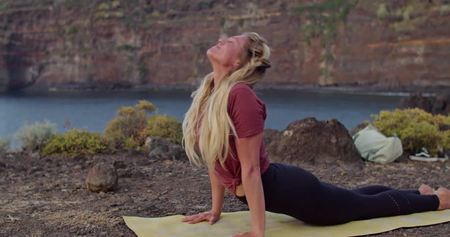 A woman stretching on a mat near a coast with water a cliff the sky and clouds in the background
