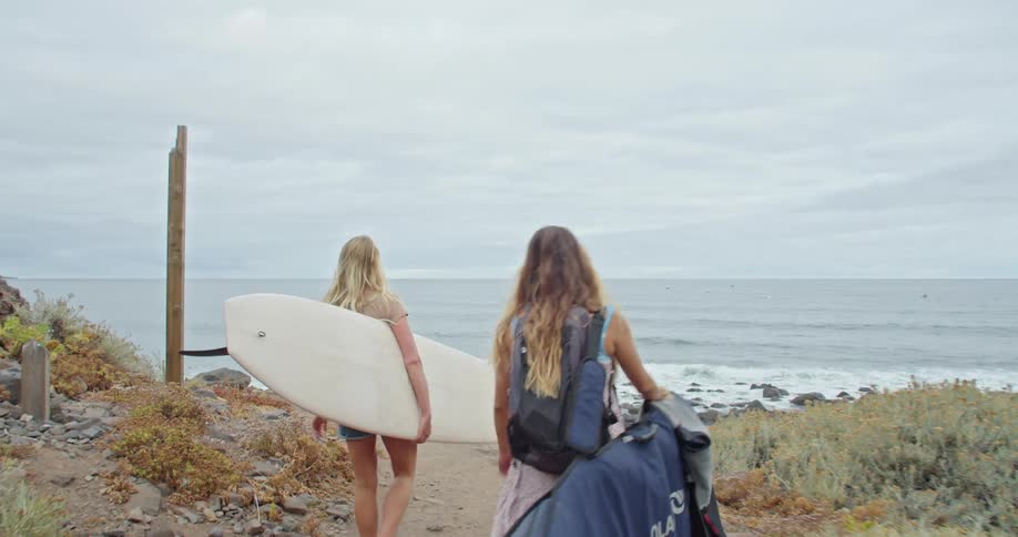 Two young women walk toward the beach with surfboards under their arms