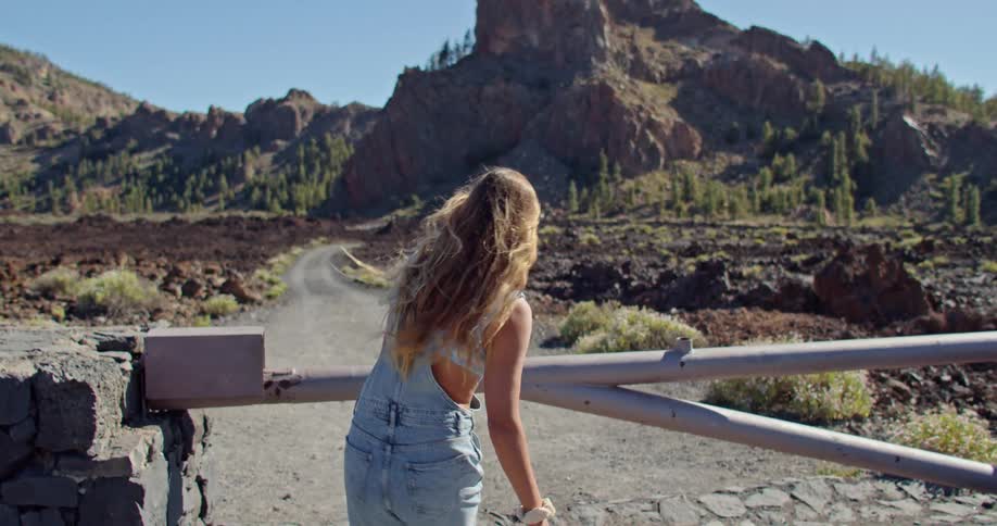 Blonde woman wearing overalls ducks under gate to walk down dirt road leading to scrub covered hill