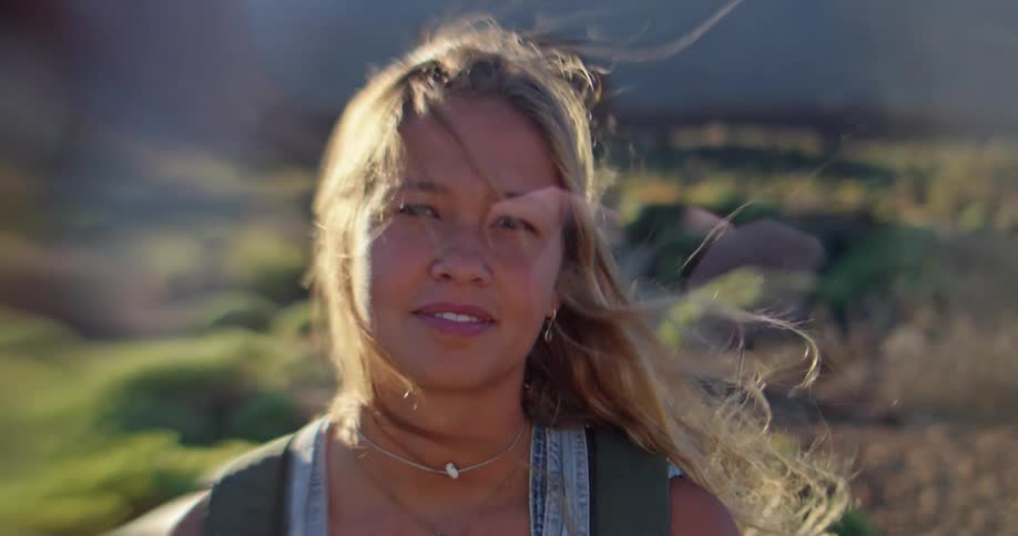 A blond woman with her hair blowing in the wind is smiling as the sun shines on her