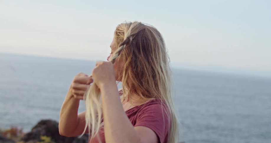 A woman near the coast braiding her hair with the ocean sky and clouds in the background