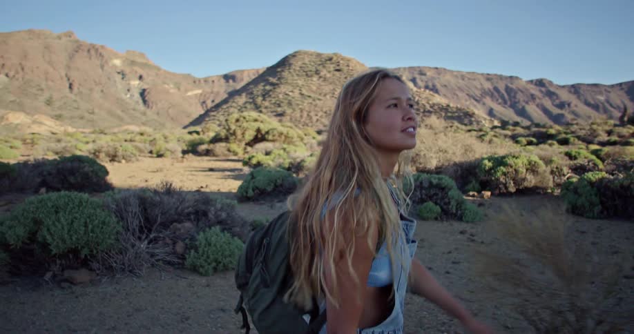 Blonde woman wearing overalls over short top and carrying backpack walks through shadowed area with scrub and hills in background under a clear blue sky