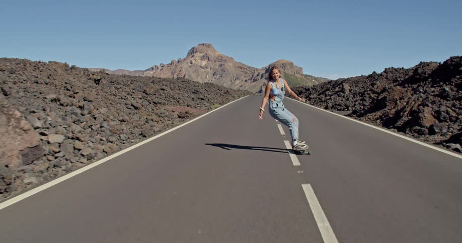 Blonde woman rides skateboard along highway through rough terrain