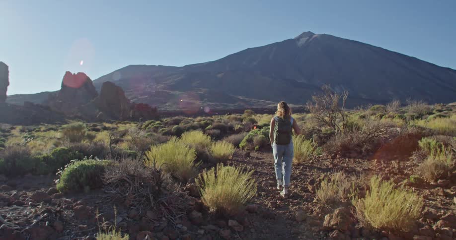 Blonde woman with strappy denim jumpsuit puts on backpack walks in the field