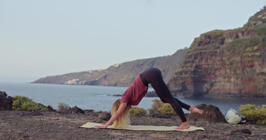 Blonde woman moves through yoga poses on yellow mat barefoot across from cliff