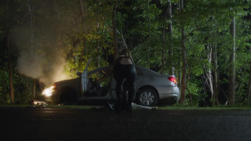 A woman in tight clothing drags an unconscious woman on the ground away from a crashed car