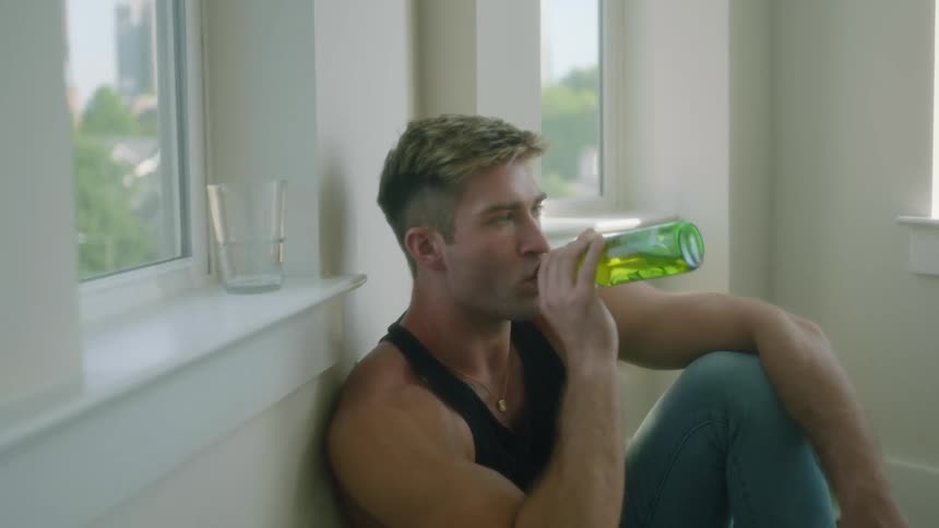 A man in a room sitting on the floor drinking a beer from a bottle with a wall and window in the background