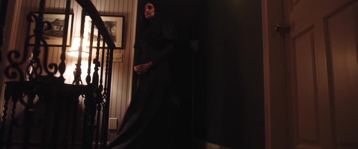 A woman in a room walking in a hallway and walking downstairs with a wall a lamp and picture frames in the background