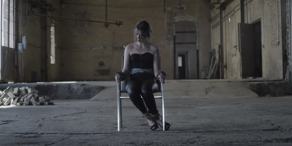 A woman bound to a chair sits in an abandoned building during the day