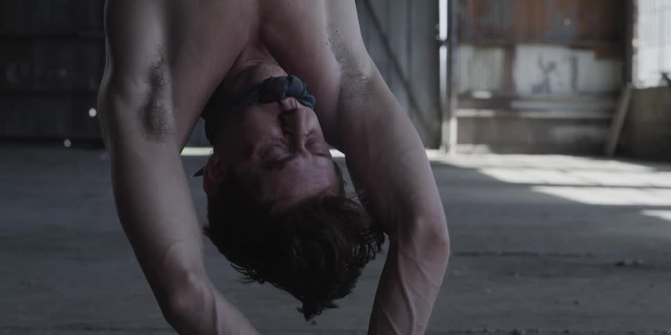 A man in a room hanging upside down with his mouth tied up with a wall in the background