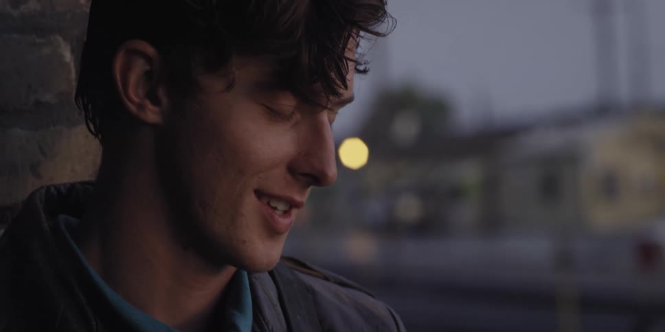 Clean shaven man with curly hair and gray coat talks to person smiling