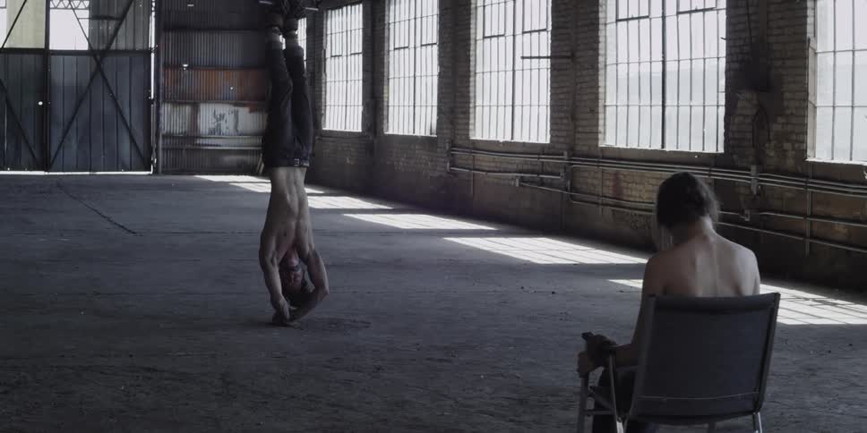 Woman tied to a chair next to a gagged man hanging upside down inside a warehouse